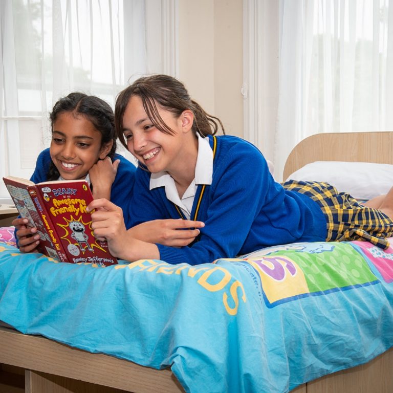 two prep school boarding students are on top of the bed in a boarding room, they are sharing a book, they look happy